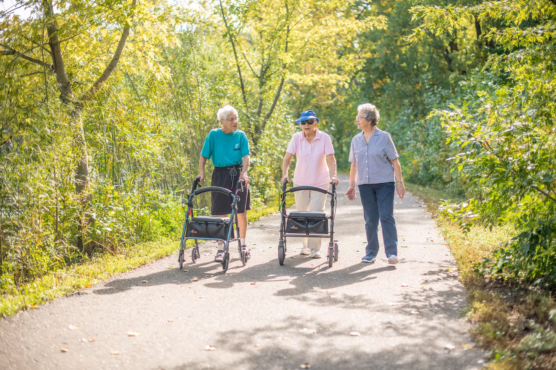 Three elderly women walking on a paved path surrounded by lush green trees and foliage. Two of the women are using walkers, and they appear to be engaged in conversation on a sunny day.