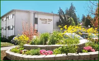 Exterior view of a two-story building with the sign 'Sunrise View Retirement, Assisted Living, Rehabilitation & Skilled Nursing' on the wall. In front of the building is a landscaped garden with various flowers and greenery, including yellow and pink blooms, surrounded by a low stone wall.