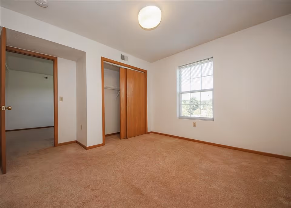 Empty carpeted bedroom with a window, an open closet with wooden sliding doors, and an open doorway to another room.