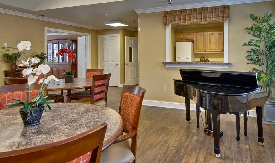 Interior view of a senior living facility common area with round tables and chairs, a black grand piano, potted plants, and a pass-through window to a kitchen area with wooden cabinets and a refrigerator.