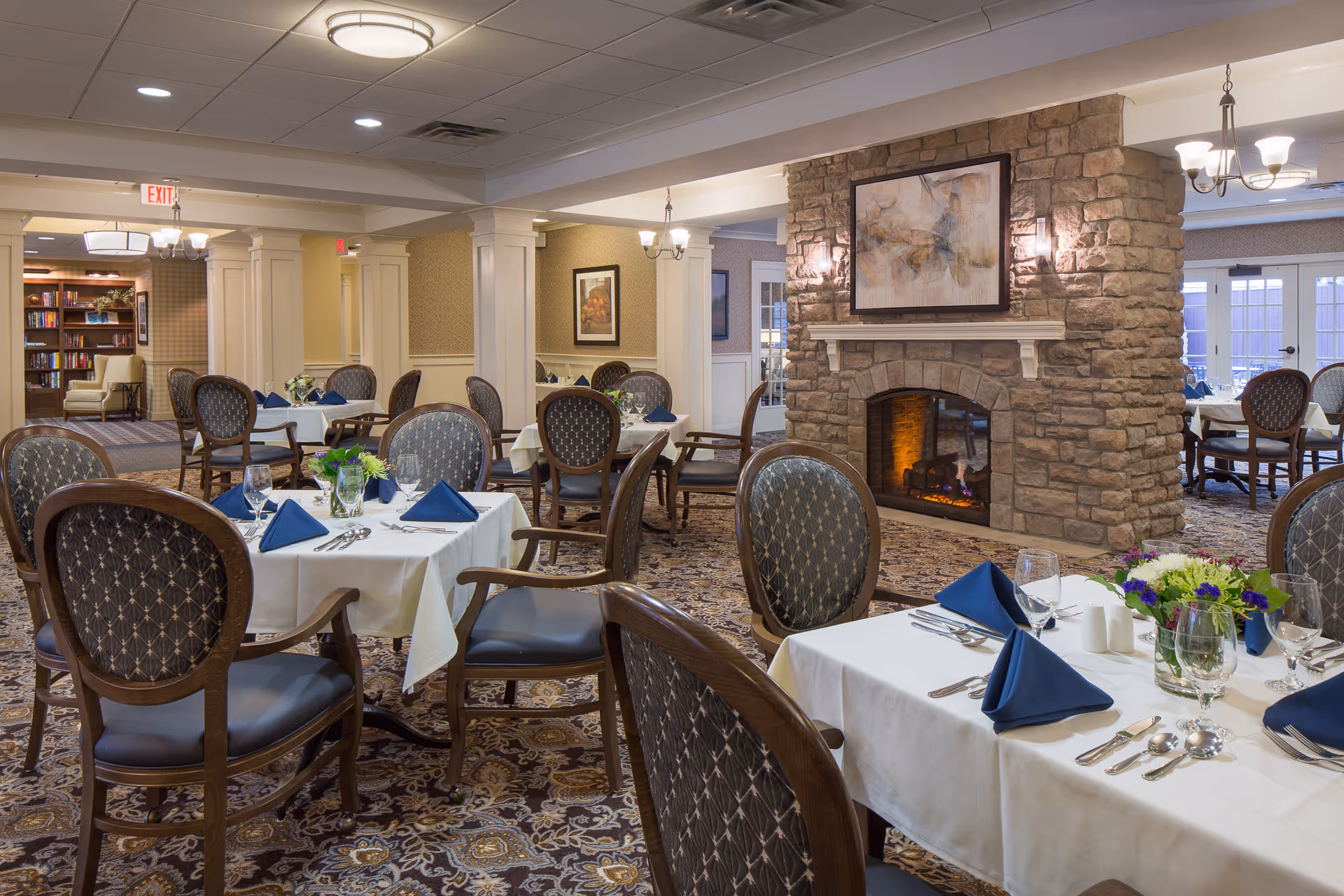 A cozy dining room in a senior living facility with tables set with white tablecloths, blue folded napkins, silverware, and glassware. The room features patterned carpet, upholstered wooden chairs, a stone fireplace with a framed abstract painting above it, and soft lighting from chandeliers and wall sconces. In the background, there is a bookshelf and additional seating areas.