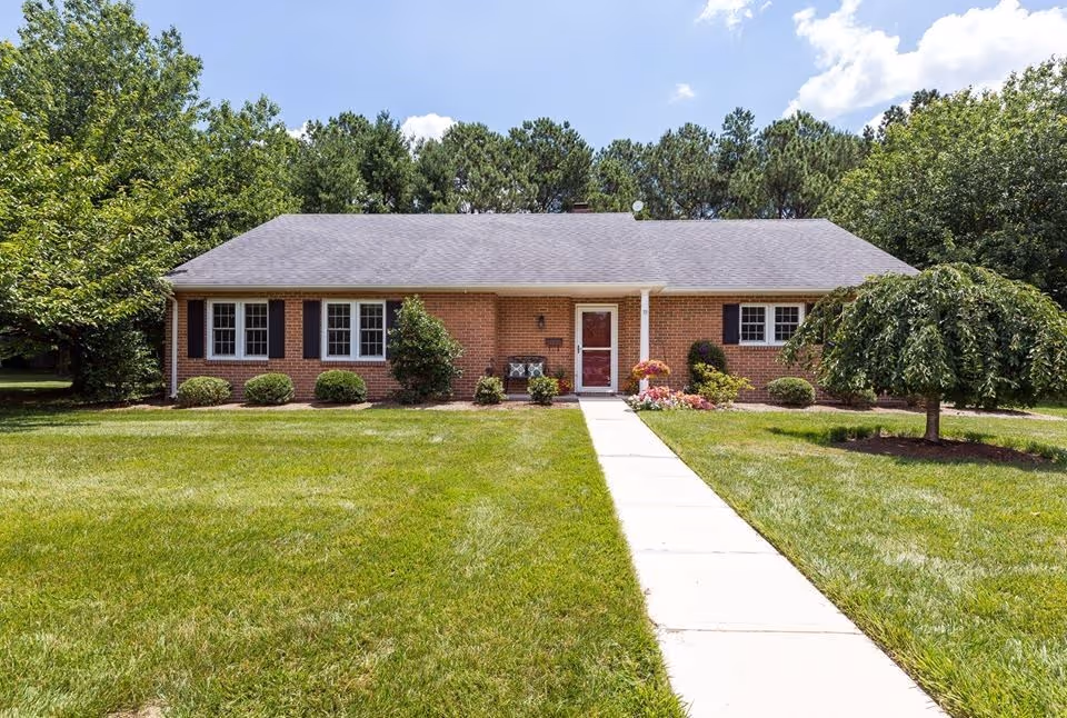 Single-story brick building with a gray shingled roof, black shutters, and white-framed windows. A concrete walkway leads to a red front door with a small porch area. The building is surrounded by a well-maintained green lawn, small bushes, and trees under a partly cloudy blue sky.