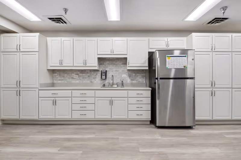 A modern kitchen with white cabinets and drawers, a stainless steel refrigerator, a sink with a faucet, and a tiled backsplash. The floor is light-colored wood, and the ceiling has fluorescent lighting panels.