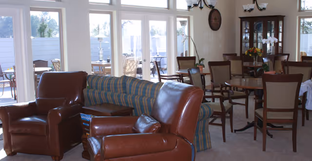 A cozy living room area with two brown leather armchairs and a striped blue sofa. Behind the seating area, there is a dining space with a round wooden table surrounded by chairs. Large windows and glass doors let in natural light and provide a view of an outdoor patio with additional seating. The room is decorated with a chandelier, a wall clock, and a glass-front cabinet with flowers on top.