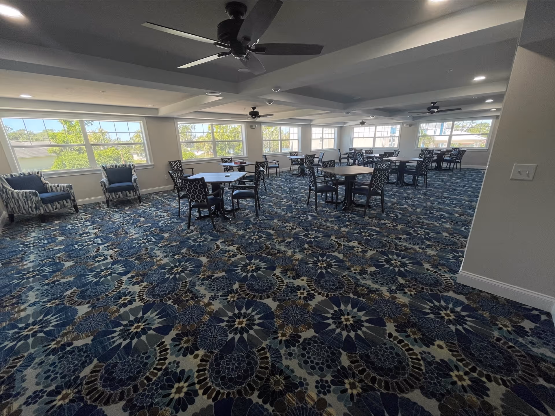 A spacious room with large windows letting in natural light, featuring multiple round tables with chairs arranged around them. The floor is covered with a blue patterned carpet, and there are several ceiling fans. A few armchairs are placed near the windows.