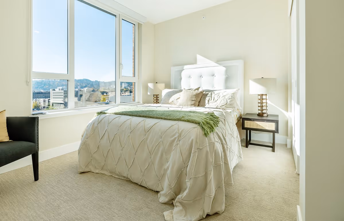 Bright bedroom with a large window showing a cityscape view. The room features a bed with a white tufted headboard, cream-colored bedding, and a green throw blanket. There are two bedside tables with matching lamps and a dark armchair with a beige pillow near the window.