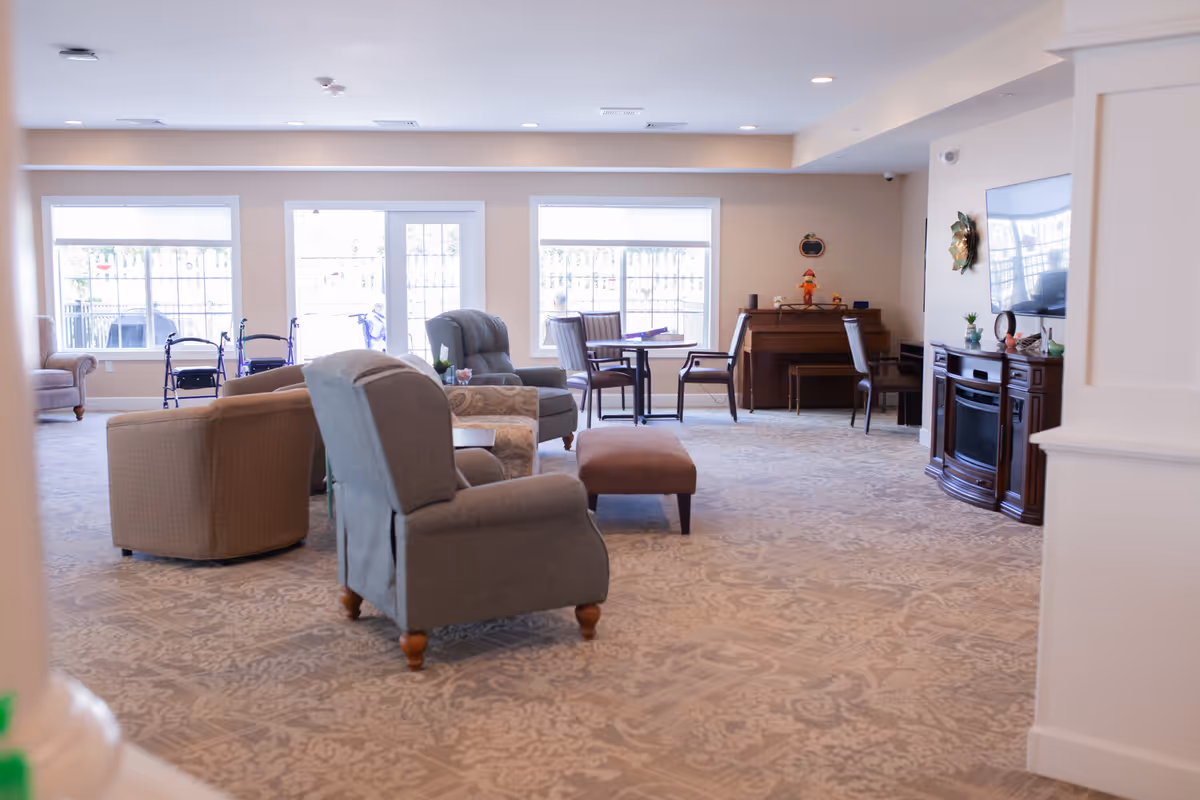 A spacious and well-lit assisted living common area with comfortable armchairs, a small table with chairs near the windows, a piano against the wall, and a flat-screen TV mounted above a wooden cabinet. The room has large windows letting in natural light and a patterned carpet covering the floor.