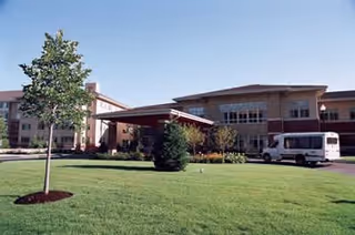 Front entrance of a multi-story senior living building with a covered driveway, green lawn, and a parked van.
