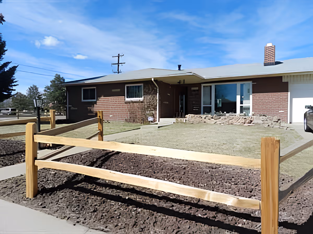 Single-story brick building with a light gray roof, a wooden split-rail fence in the foreground, and a patchy lawn. The building has a large window and a garage door on the right side. Trees and a clear blue sky are visible in the background.