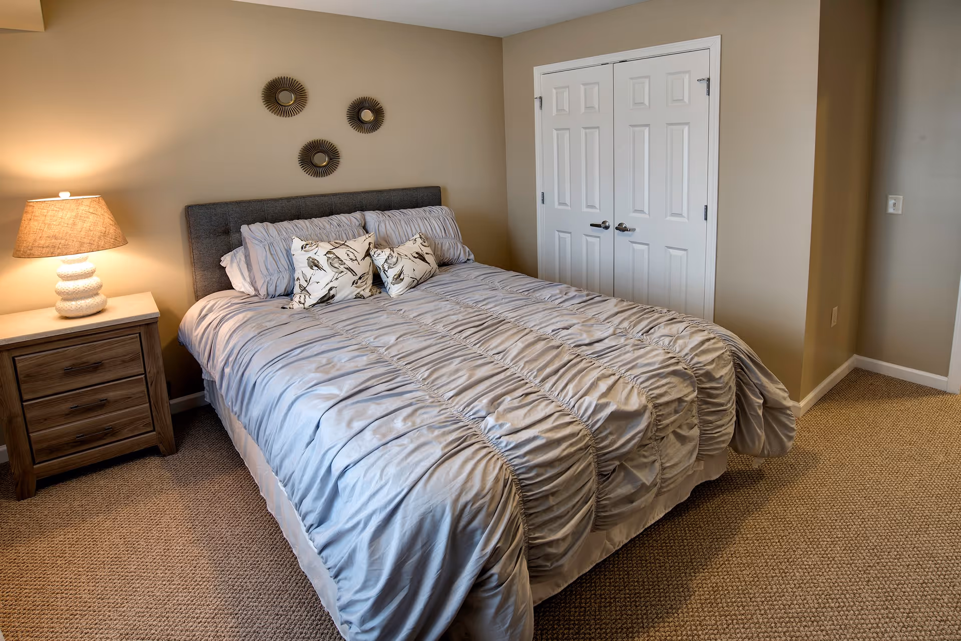 A neatly made bed with a gray ruched comforter and decorative pillows featuring bird designs in a bedroom. There is a wooden nightstand with a lamp on the left side of the bed. The walls are beige, and there are three decorative round wall hangings above the bed. A white double-door closet is visible on the right side of the room.