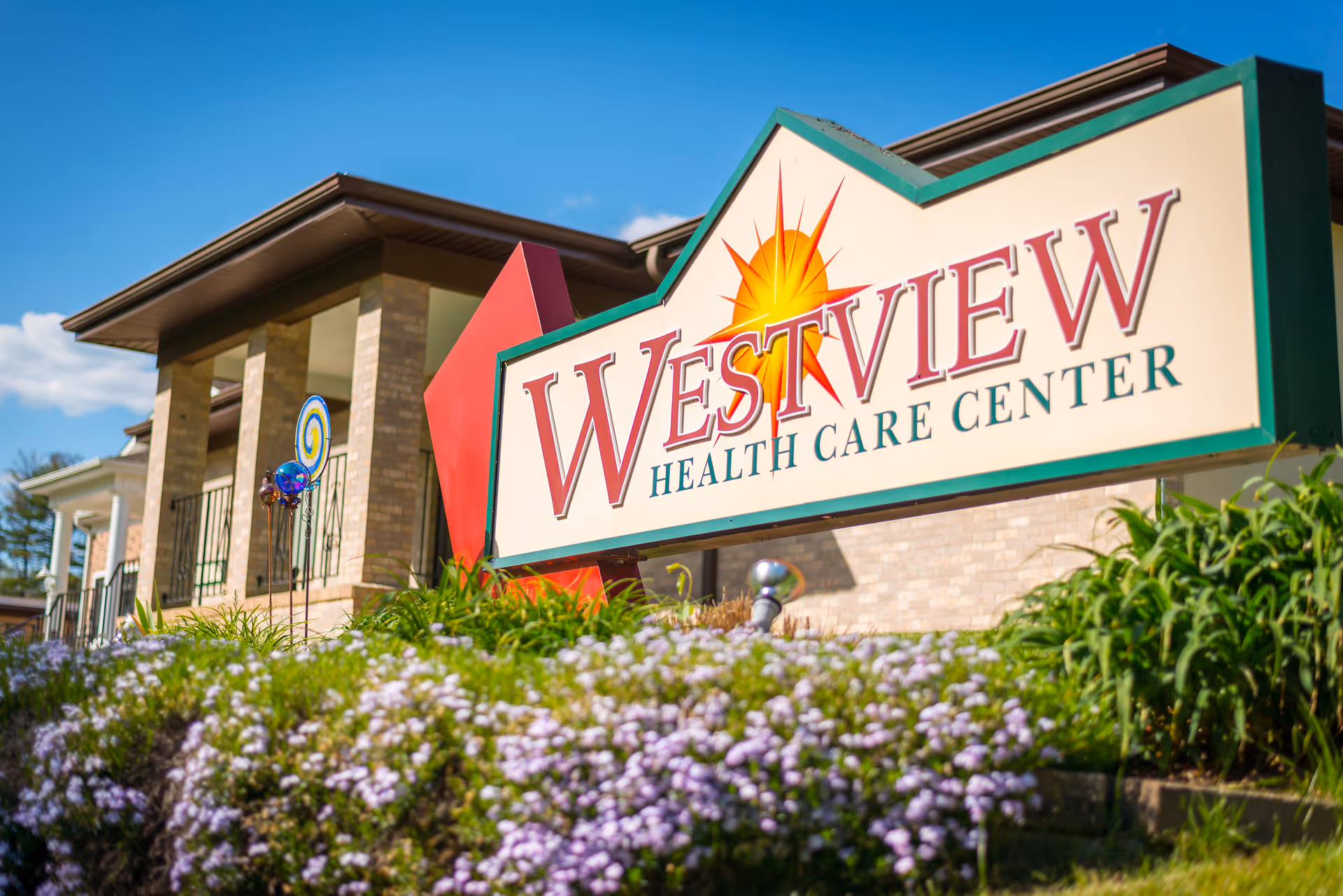 Outdoor view of the Westview Health Care Center sign with a building and garden featuring purple flowers in the background under a clear blue sky.