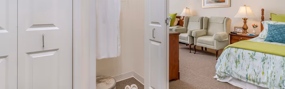 View of a senior living facility bedroom with a bed featuring green and white bedding, two beige armchairs, a wooden nightstand with a lamp, and a framed picture on the wall. The image is taken from a hallway showing white closet doors partially open.