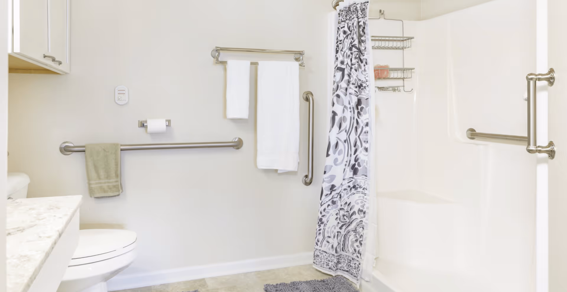 A clean and accessible bathroom featuring a white toilet, a marble countertop, a shower with a patterned curtain, grab bars on the walls, and towels hanging on towel racks.