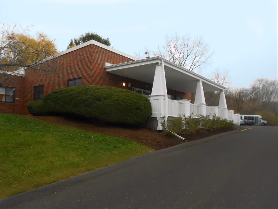Exterior view of a single-story brick building with a white covered porch supported by large white columns. There is a neatly trimmed green bush in front of the building and a paved driveway or parking area to the right. Trees with bare branches are visible in the background under a cloudy sky.