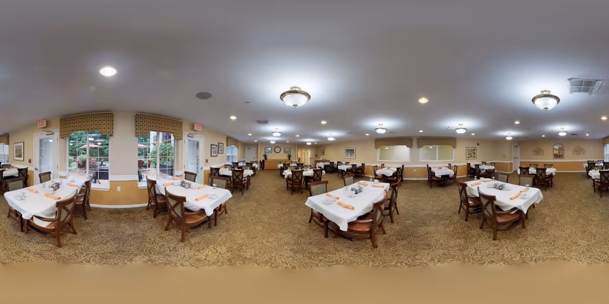 A spacious dining room with multiple tables covered in white tablecloths and set with napkins and silverware. The room has beige walls, patterned carpet, and several ceiling lights. Large windows and glass doors provide a view of an outdoor garden area with greenery and patio umbrellas.