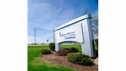 Outdoor view of a large white sign with blue text reading 'Signature HealthCARE of Bluffton' situated on a landscaped area with green grass, bushes, and a clear blue sky in the background.