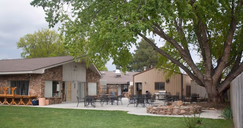 Outdoor patio area at a senior living facility with several black metal tables and chairs arranged on a concrete surface. There is a large tree providing shade, a stone fire pit, and buildings with stone and beige siding in the background. Green grass surrounds the patio area.
