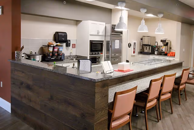 Interior view of a senior living facility kitchen area with a long counter and several chairs lined up. The counter has a granite surface and is equipped with various kitchen appliances including a coffee maker, oven, and a beverage dispenser. The walls are painted in neutral tones and there are three pendant lights hanging above the counter.