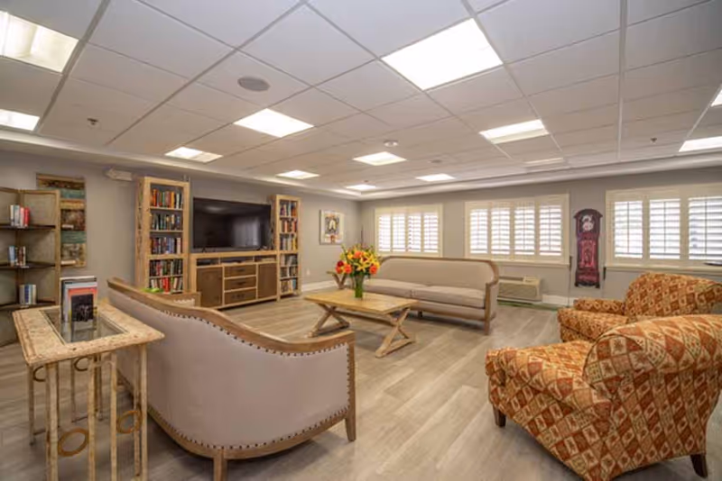 Well-lit communal lounge with sofas, patterned armchairs, a coffee table, TV and bookshelves.