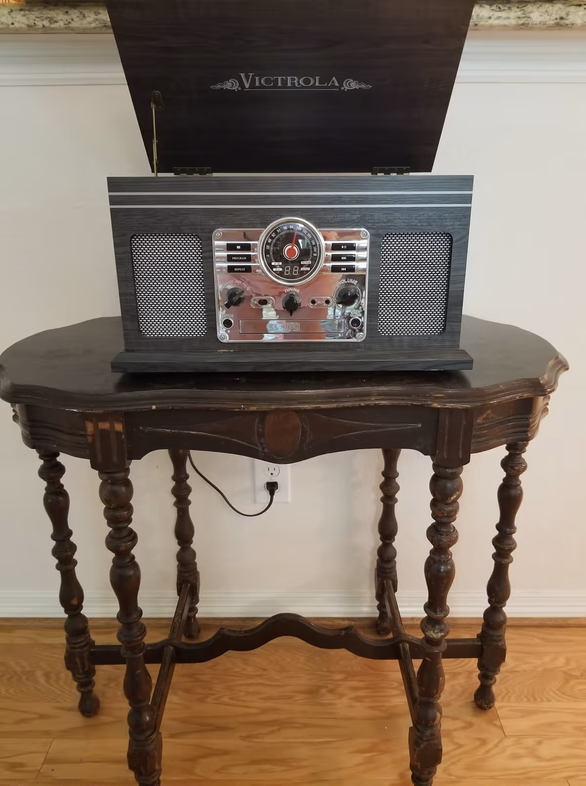 A vintage-style Victrola record player with an open lid, placed on an ornate wooden table with turned legs. The table is set against a white wall with a power outlet and a plugged-in black cord visible behind it. The floor is wooden.