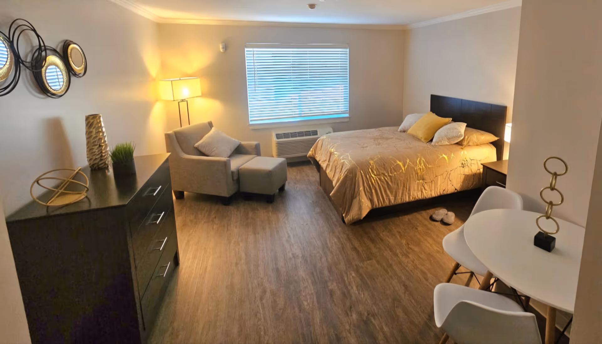 Well-lit studio-style bedroom with a bed, armchair and ottoman, dresser, small dining table, and a window with blinds.