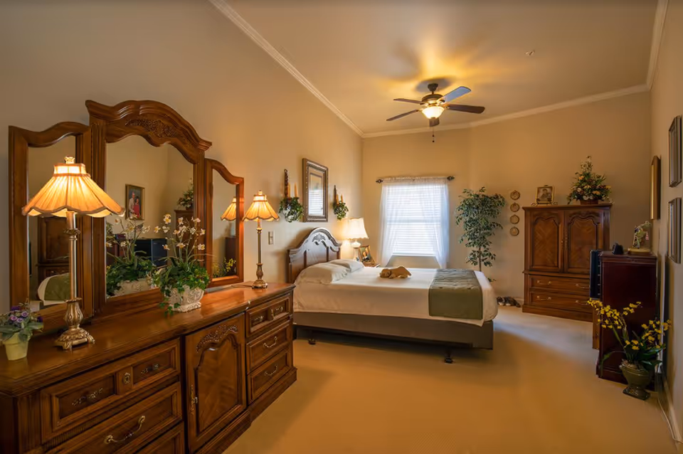 A warmly lit bedroom with beige walls and carpet, featuring a large wooden dresser with three mirrors and two lamps, a neatly made bed with white linens and a green throw, a ceiling fan with light, a window with sheer white curtains, a wooden armoire, and various plants and framed pictures decorating the room.