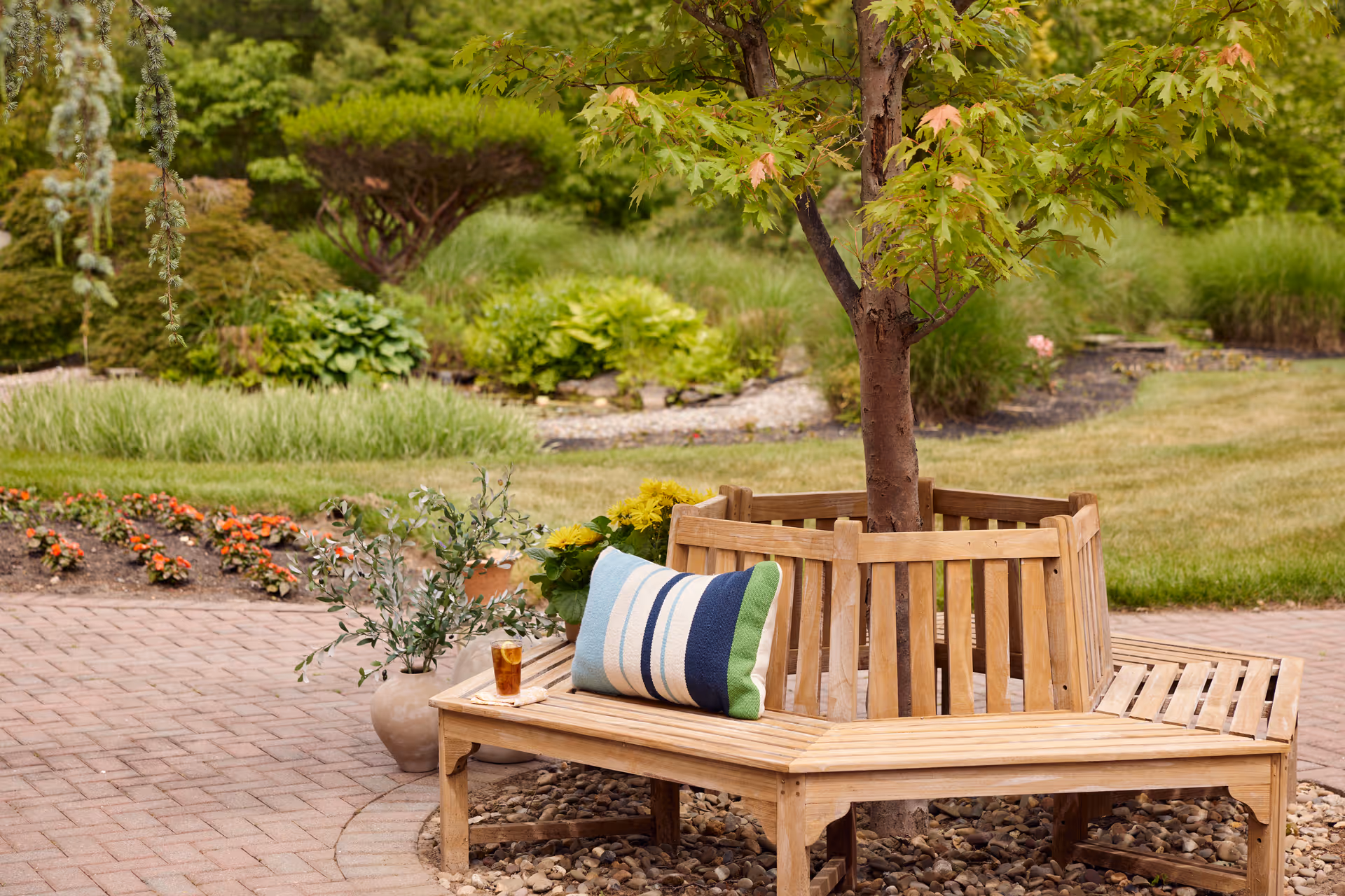 A wooden circular bench surrounds a tree in a landscaped garden area with green shrubs, grass, and flower beds. A striped pillow and a glass of iced tea are placed on the bench, with a potted plant nearby on a brick-paved patio.