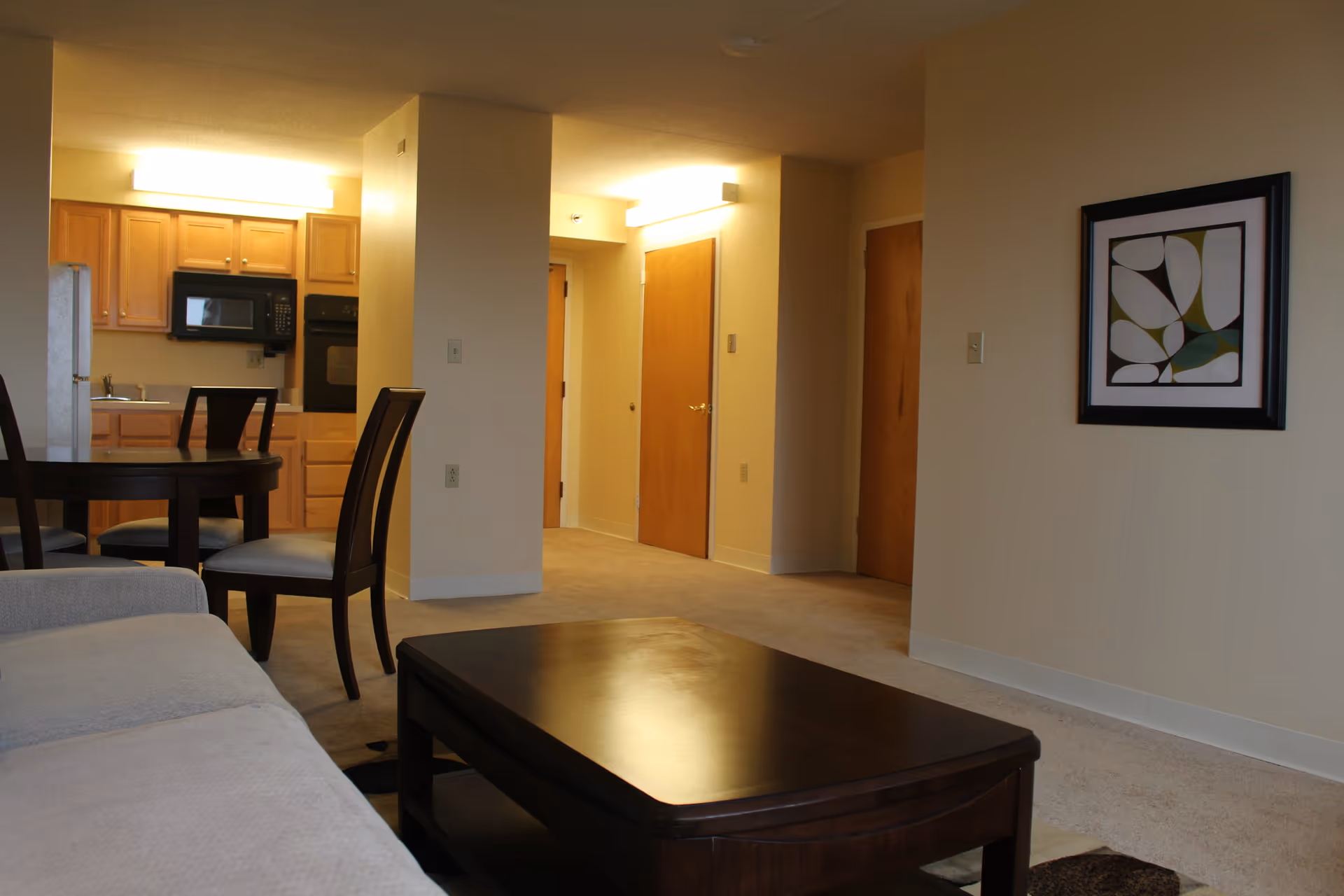 Interior view of a senior living facility apartment showing a beige couch in the foreground, a dark wooden coffee table, a dining table with chairs, a kitchen area with wooden cabinets, a microwave, and an oven. The walls are light-colored with a framed abstract artwork hanging on one wall. There are multiple wooden doors visible in the hallway area.