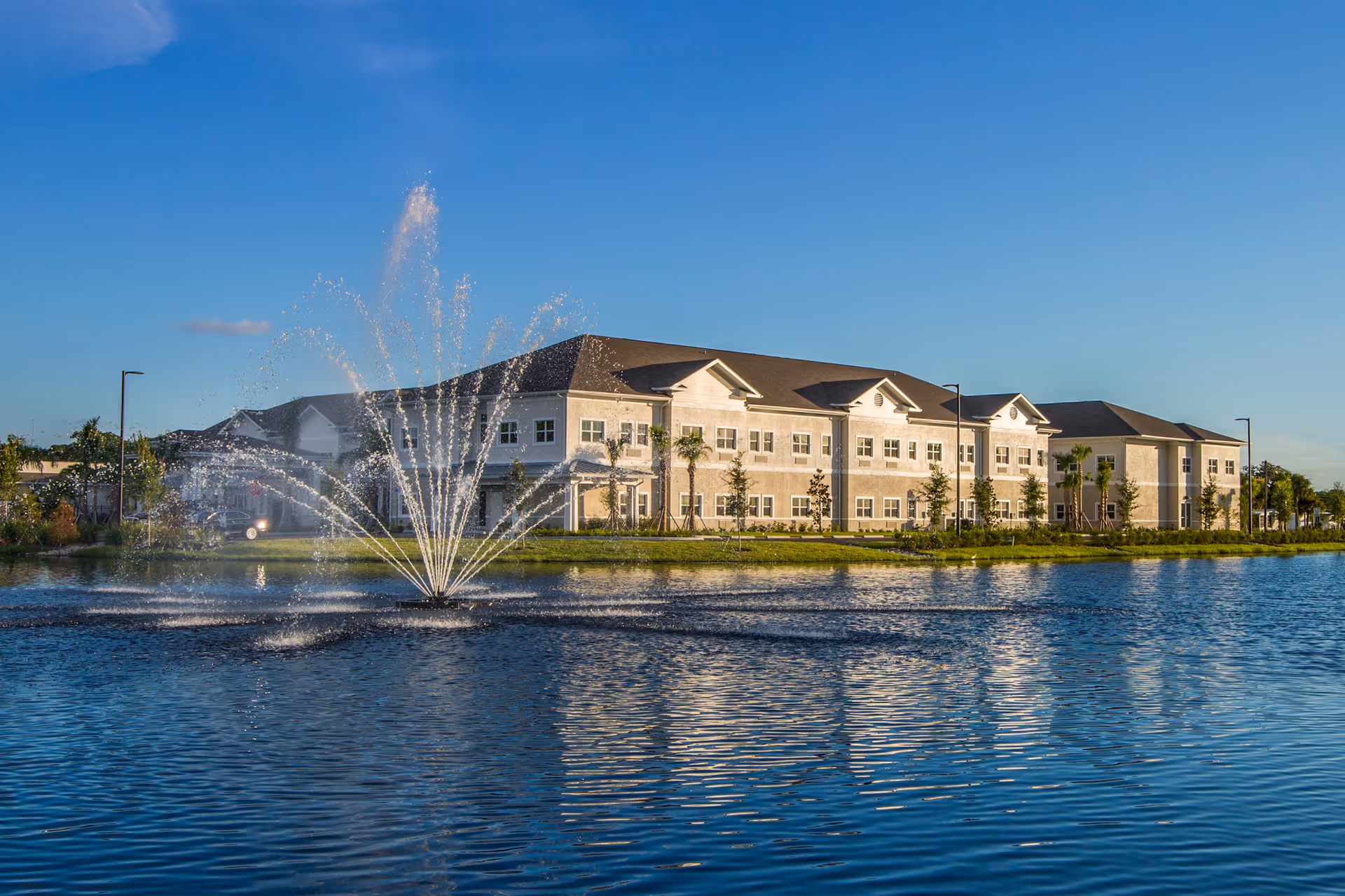 A large two-story building with multiple windows situated behind a pond with a water fountain spraying water into the air, under a clear blue sky.
