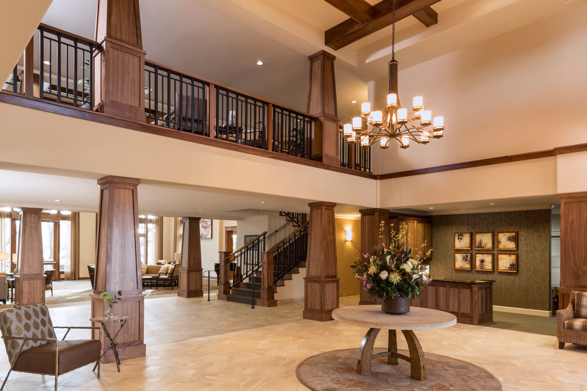 Spacious and elegant senior living facility lobby with wooden pillars, a round table with a large floral arrangement in the center, a chandelier hanging from the ceiling, and a staircase leading to an upper floor with a railing. Comfortable seating areas are visible in the background.