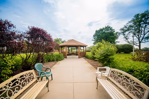 Outdoor garden area with a paved walkway leading to a wooden gazebo. The walkway is flanked by two decorative benches and a plastic chair, surrounded by green bushes, trees, and plants under a partly cloudy sky.