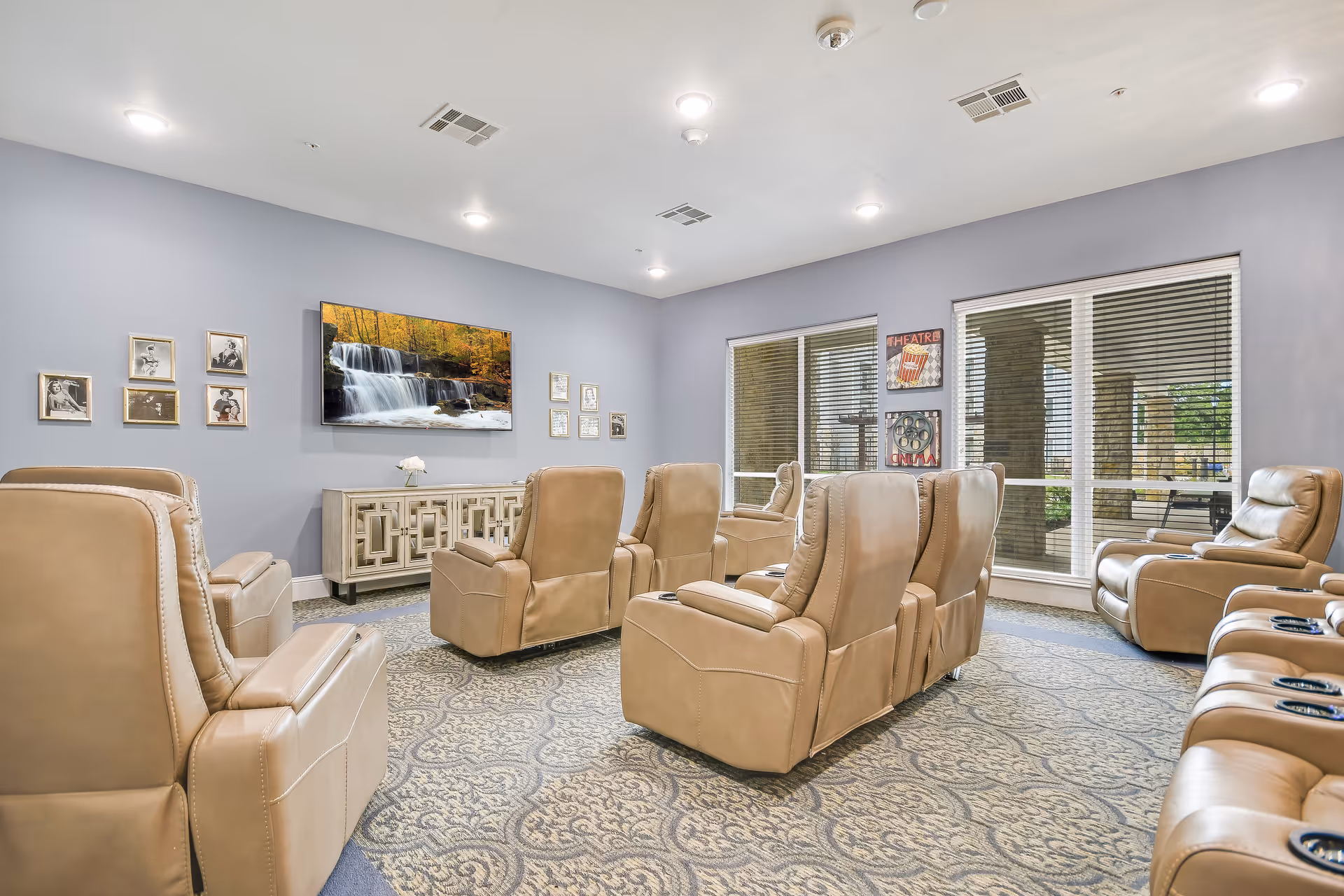 A comfortable senior living media room with beige recliner chairs arranged facing a wall-mounted TV and large windows.