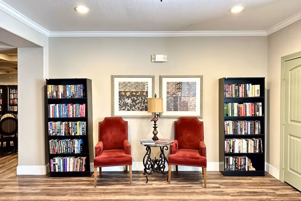 Two red upholstered chairs flank a small decorative table and lamp between two bookcases in a bright interior reading nook.