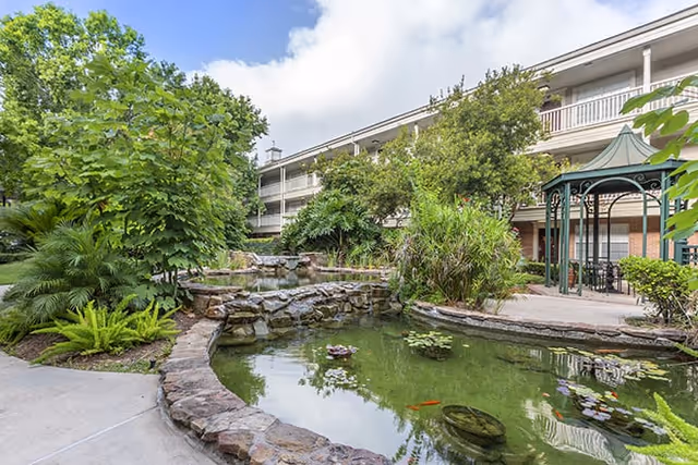 Outdoor garden area at Brookdale West University featuring a small pond with lily pads and a few fish, surrounded by lush greenery and trees. A stone border edges the pond, and a gazebo with seating is visible on the right side. The multi-story building with balconies overlooks the garden under a partly cloudy sky.