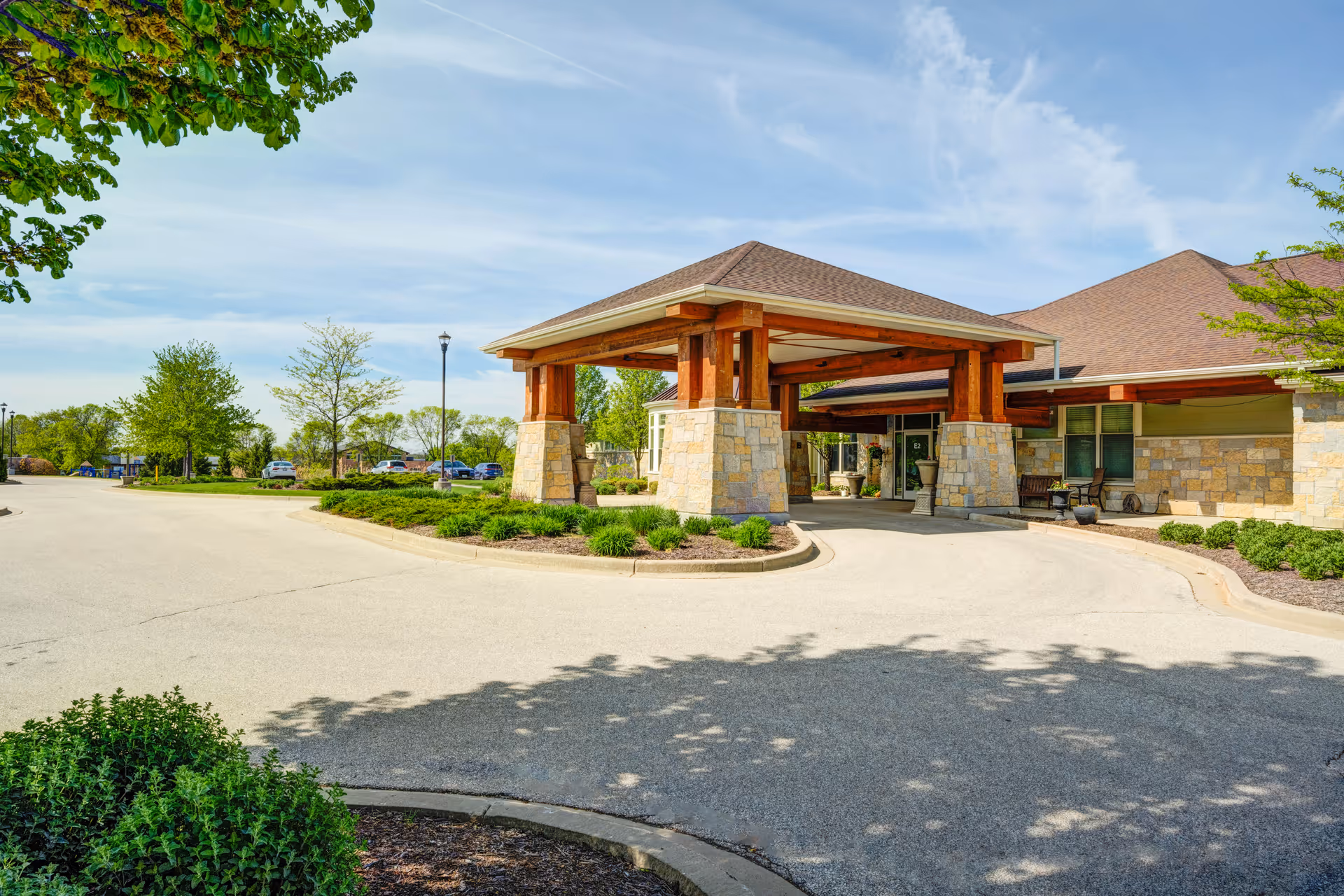 Front entrance canopy of a senior living facility with stone pillars, a circular driveway, and landscaped grounds under a blue sky.