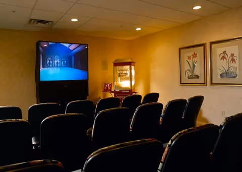 A small theater room with several rows of black cushioned chairs facing a large screen displaying an image of an indoor pool area. To the right of the screen is a popcorn machine and two framed floral paintings on the wall. The room has a beige ceiling with recessed lighting and beige walls.