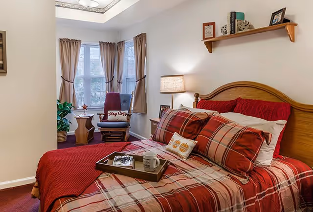 A cozy bedroom with a wooden bed frame and red plaid bedding. A tray with a coffee mug and a book rests on the bed. There is a wooden rocking chair with a red cushion and a pillow that says 'Stay Cozy' near a window with beige curtains. A small side table with a plant and a lamp on a nightstand beside the bed are also visible. A wooden shelf with books and decorative items is mounted on the wall above the bed.