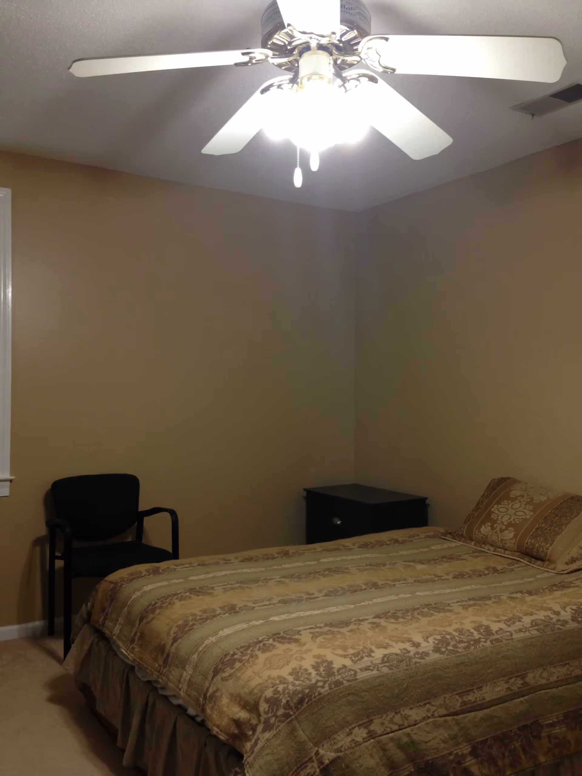 A bedroom with beige walls and carpeted floor, featuring a bed with a patterned bedspread and matching pillow. There is a black chair near the window and a small black nightstand next to the bed. A ceiling fan with lights is turned on above the bed.