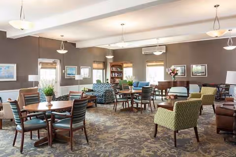 Spacious senior living lounge with assorted seating, tables, bookshelves, and plants.