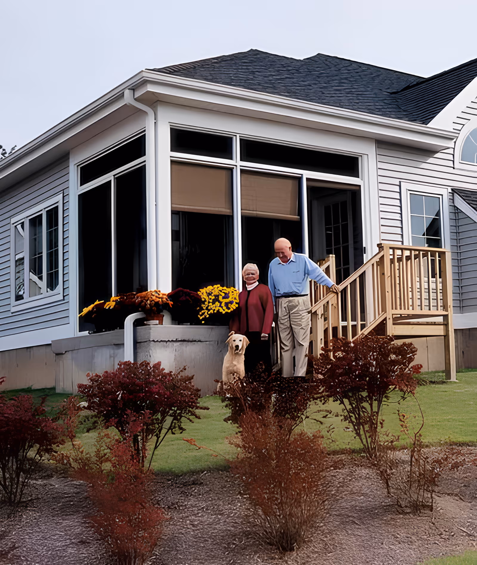 Two elderly people and a dog standing on the lawn in front of a single-story house with large windows and a small wooden porch.