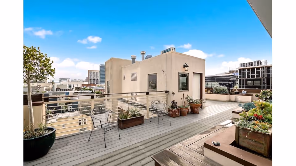 A spacious rooftop patio with wooden decking, metal chairs, and various potted plants including succulents and small trees. The patio is surrounded by a metal railing and overlooks a cityscape with buildings under a blue sky with scattered clouds.