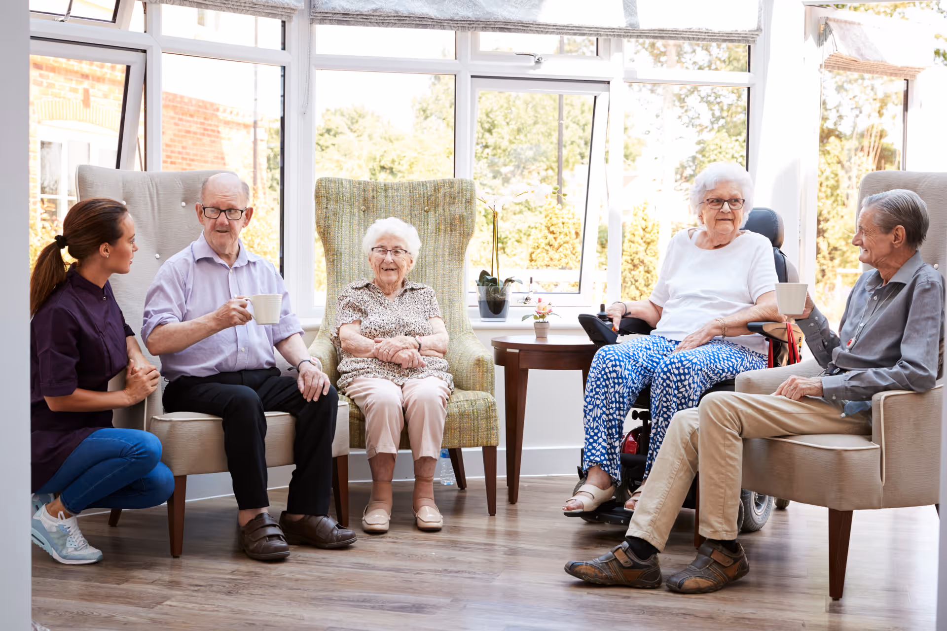 A caregiver kneels and chats with four elderly residents seated in a bright communal living room.