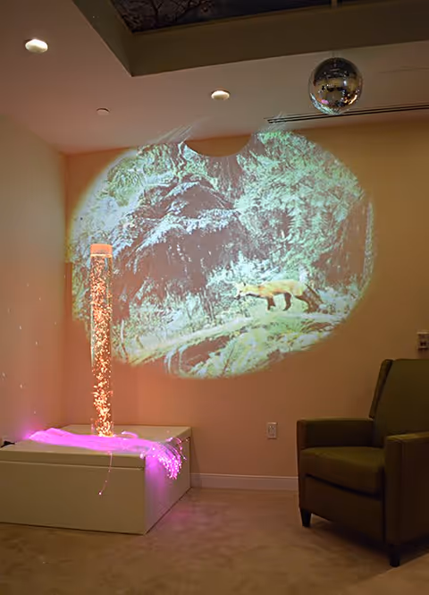 A cozy room corner with a green armchair and a sensory bubble tube light emitting pink light. A nature scene with a fox in a forest is projected on the beige wall above the bubble tube. A disco ball hangs from the ceiling, reflecting light around the room.