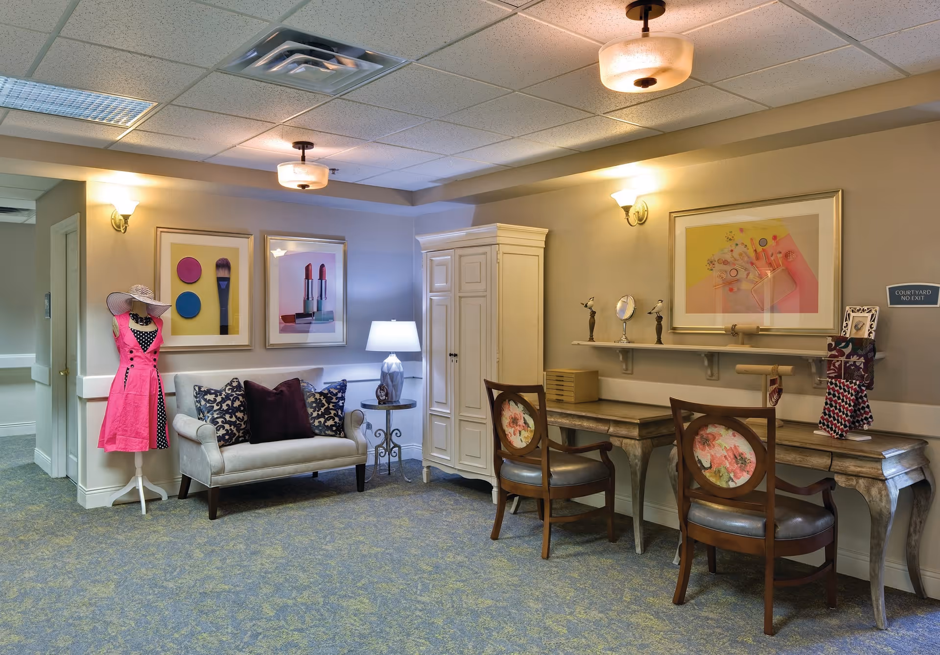 Comfortable sitting and vanity area with a sofa, two decorative chairs at a desk, a mannequin in a pink dress, and framed artwork on the walls.