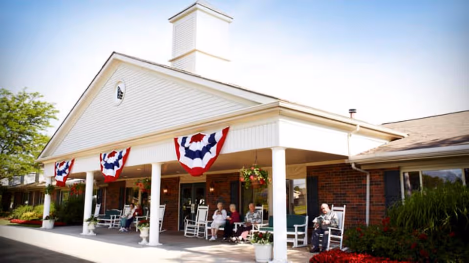 Front porch of a senior living facility decorated with red, white, and blue bunting. Several elderly residents are sitting on rocking chairs under the covered porch, enjoying the outdoor space. The building has white columns and a brick exterior with hanging flower baskets.