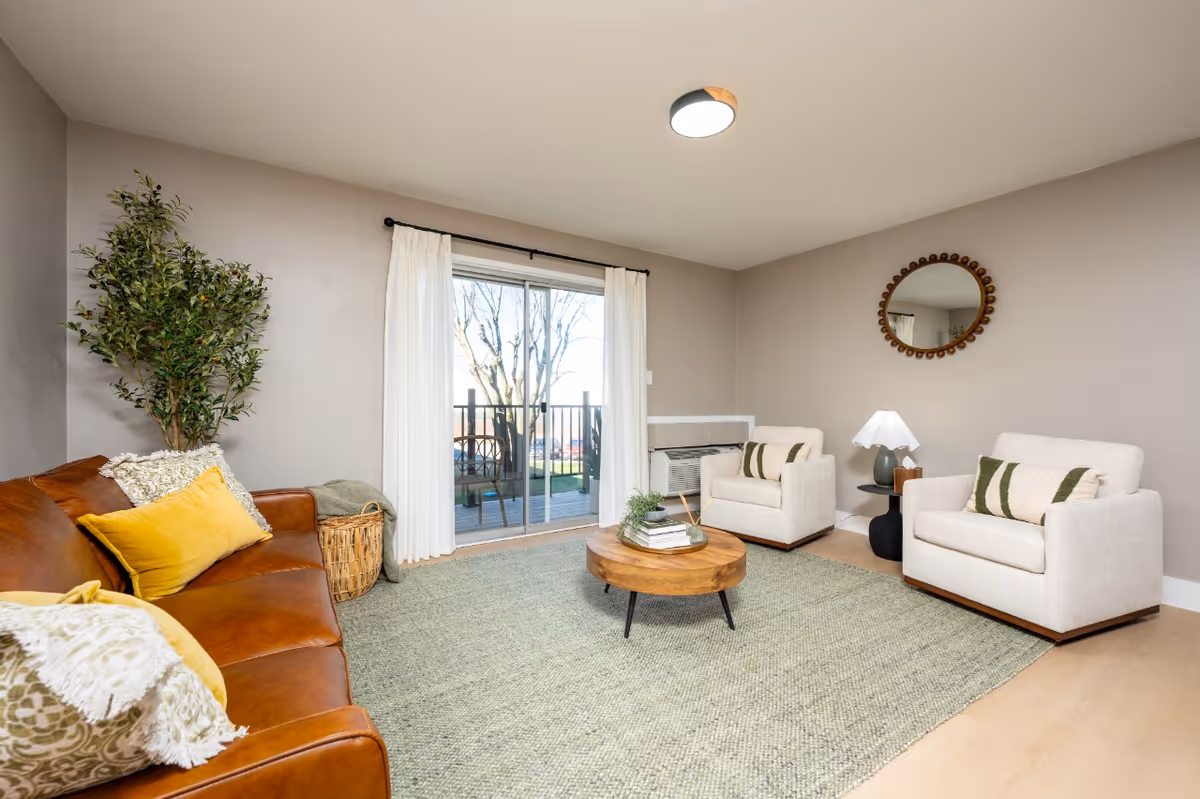 A cozy living room with a brown leather sofa adorned with yellow and patterned pillows on the left, two beige armchairs with green and beige pillows on the right, a round wooden coffee table with books and a small plant in the center, a large green rug covering the floor, a round mirror on the wall above the armchairs, a small black side table with a lamp, a large potted plant in the corner, and sliding glass doors with white curtains leading to an outdoor balcony.
