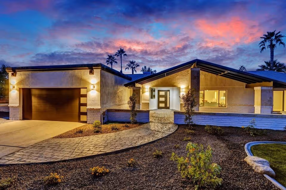 Exterior view of a modern single-story house at dusk with warm lights illuminating the entrance and garage. The sky is colorful with shades of pink and purple, and there are palm trees in the background. The front yard has a paved walkway and landscaped plants.