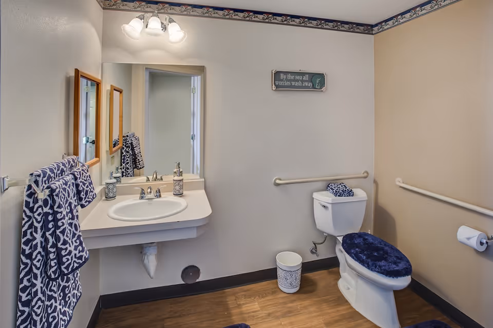 A clean and accessible bathroom with beige walls and wood-look flooring. The room features a white toilet with a dark blue fuzzy seat cover and matching towel on top. There are grab bars on the walls near the toilet and a toilet paper holder. A white sink with a countertop is mounted on the wall, with a large mirror above it and two decorative soap dispensers. Blue patterned towels hang on a towel rack next to the sink. Above the toilet, a small decorative sign reads 'By the sea all worries wash away.' The bathroom has a nautical-themed border near the ceiling.