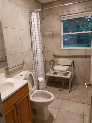 A bathroom with beige tiled walls and floor, featuring a toilet with the lid open, a wooden vanity with a sink, a shower area with a white and gray striped curtain, a white shower chair, grab bars on the walls, and a window showing greenery outside.