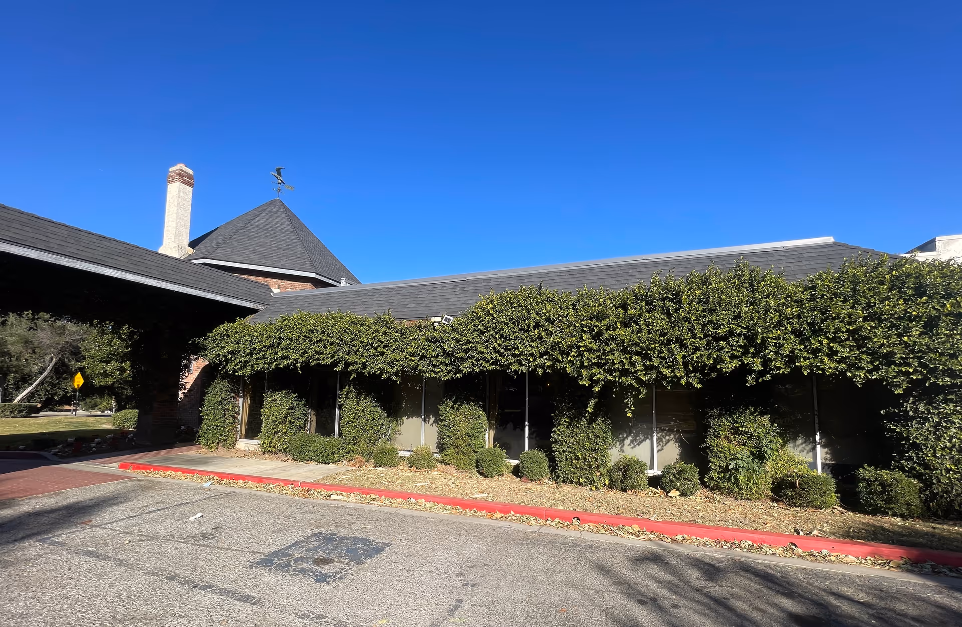 Exterior view of a building with a sloped roof and chimney, partially covered with green ivy and bushes along the front. The sky is clear and blue, and there is a red curb along the edge of the parking area in front of the building.